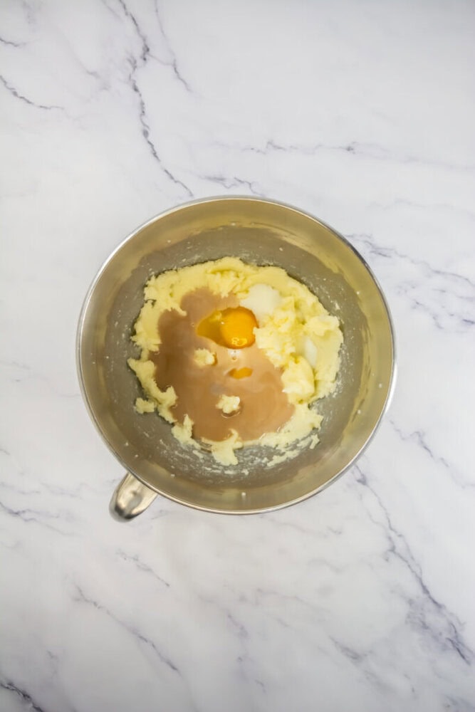 A metal mixing bowl contains butter, sugar, vanilla extract, and an egg on a white marble countertop, ready to be mixed.