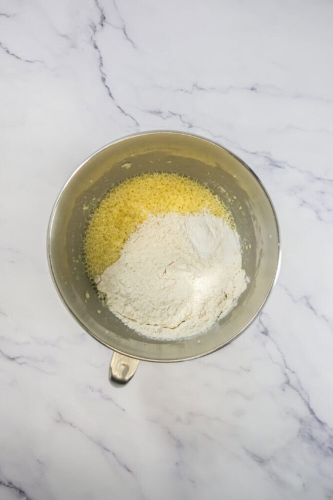 A metal mixing bowl containing creamed wet ingredients and a mound of flour on top, placed on a white marble surface.