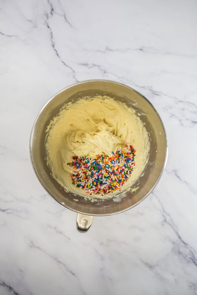 A metal mixing bowl containing cookie dough with colorful sprinkles on a white marble surface.