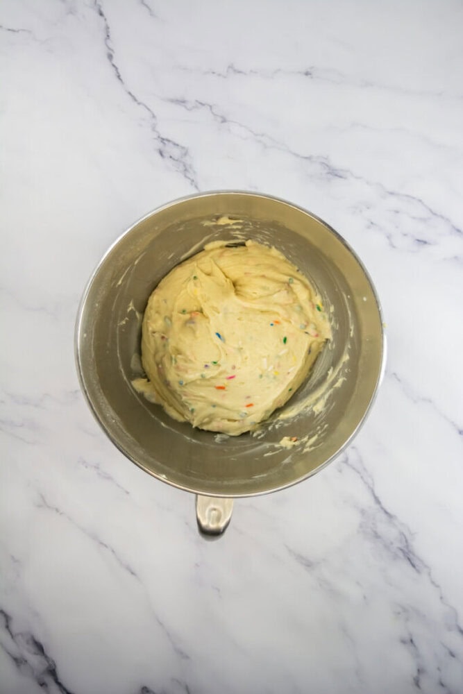 A metal mixing bowl containing cookie dough with colorful sprinkles, placed on a white marble surface.