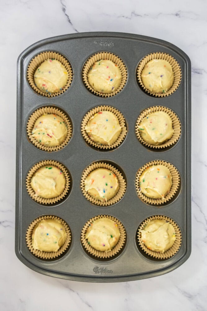 A muffin tin with twelve cupcake liners filled with uncooked batter containing colorful sprinkles, placed on a white marble surface.