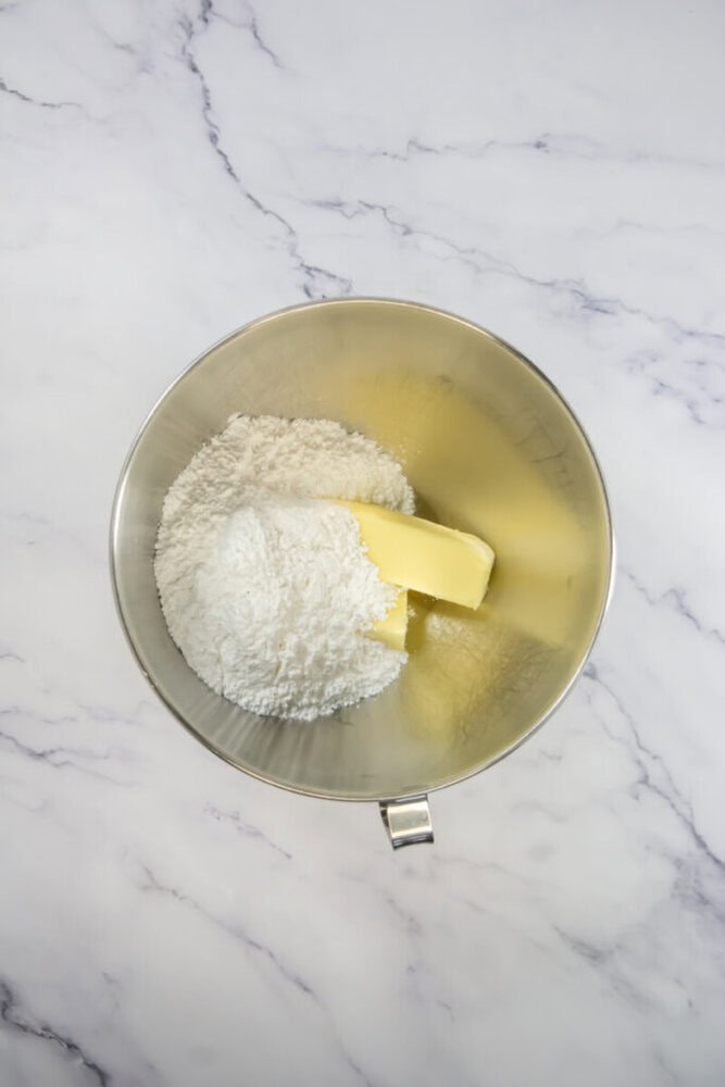 A metal mixing bowl containing powdered sugar and two sticks of butter, placed on a white marble surface.