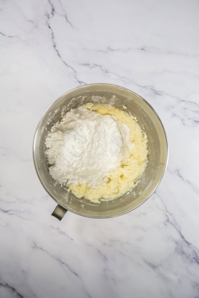 A metal mixing bowl containing creamed butter and sugar with flour added on top, placed on a white marble surface.