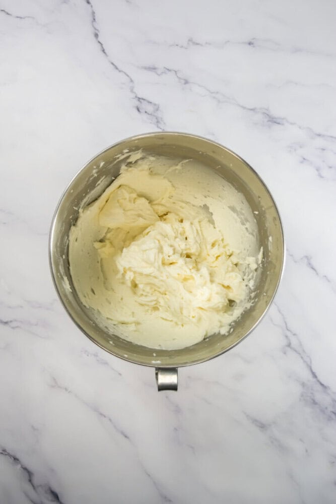 A metal mixing bowl filled with whipped cream cheese frosting sits on a white marble countertop.