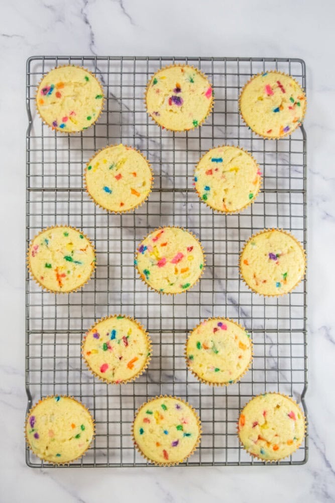 Twelve funfetti cupcakes with colorful sprinkles on top are arranged on a cooling rack on a white marble surface.