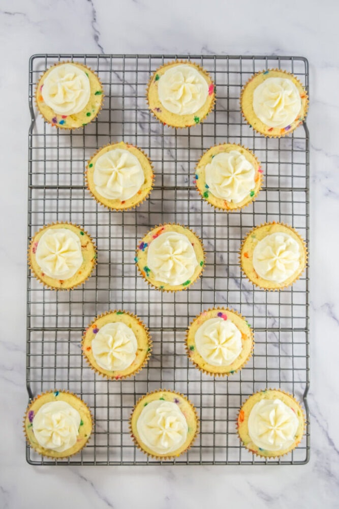 A cooling rack with twelve cupcakes topped with white swirled frosting and colorful sprinkles, arranged on a marble surface.
