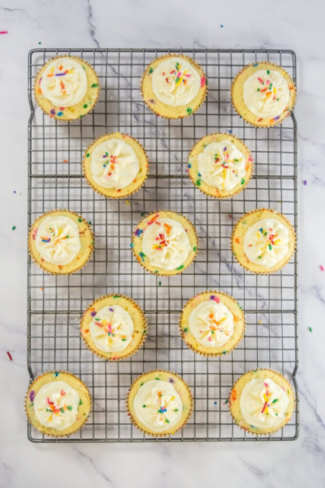 A cooling rack with twelve cupcakes topped with white frosting and colorful sprinkles on a marble surface.