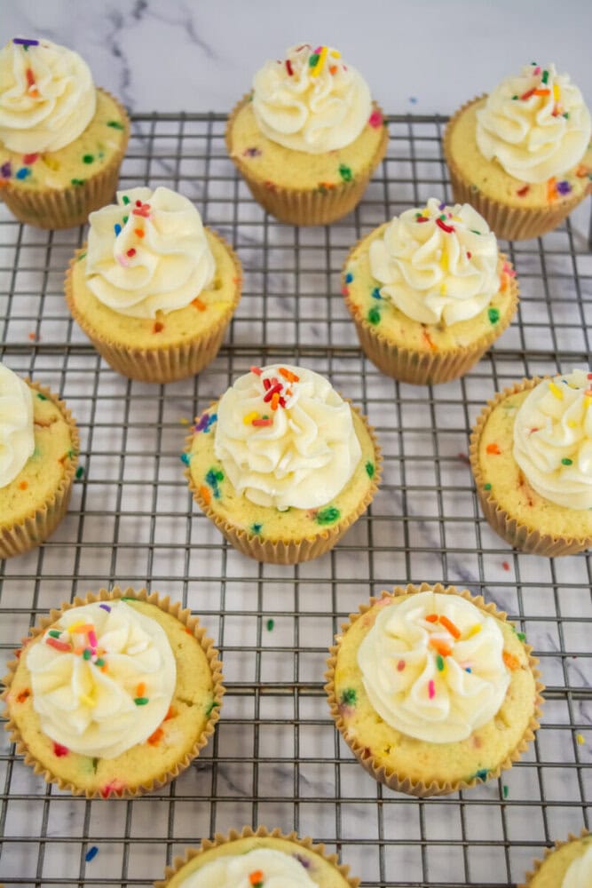Twelve cupcakes with white frosting and colorful sprinkles are arranged on a cooling rack over a marble surface.