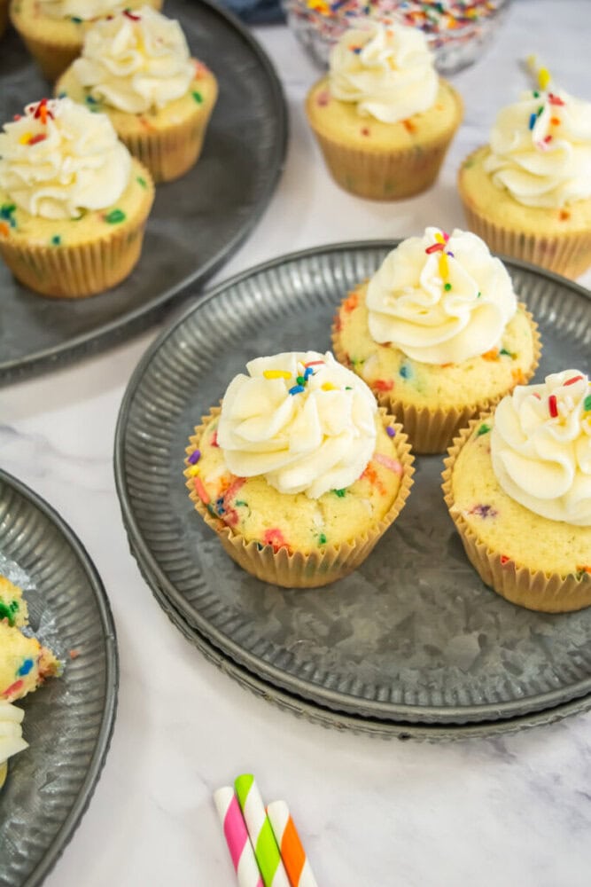 Four cupcakes with white frosting and colorful sprinkles sit on a metal plate, surrounded by more cupcakes on additional plates. Three striped paper straws are placed nearby.