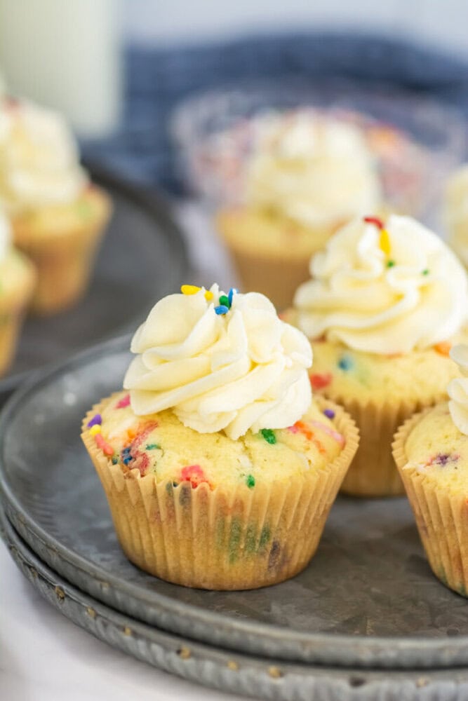 A close-up of funfetti cupcakes with white frosting and colorful sprinkles, arranged on a gray plate.