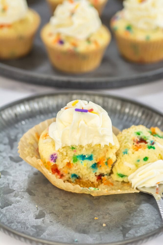 A cupcake with colorful sprinkles and white frosting is cut open on a plate, with more similar cupcakes on a tray in the background.