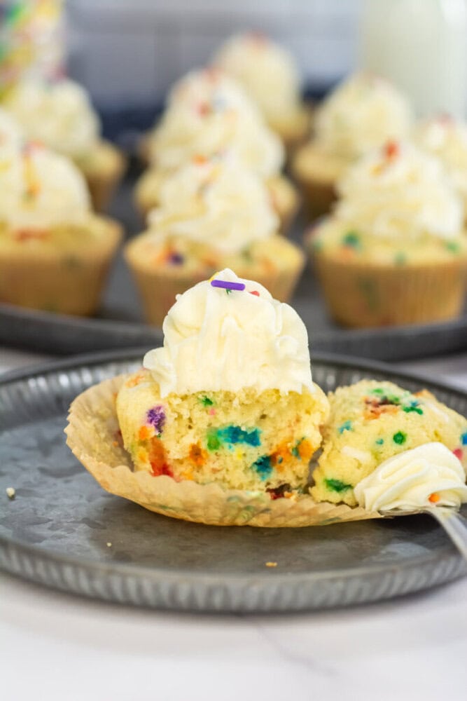 A partially eaten funfetti cupcake with white frosting sits on a gray plate in front of more frosted cupcakes.