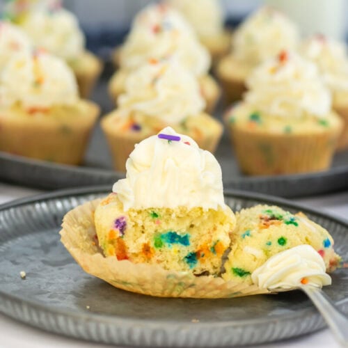 A plate with a funfetti cupcake cut in half, topped with swirled white frosting and sprinkles, surrounded by more cupcakes in the background.