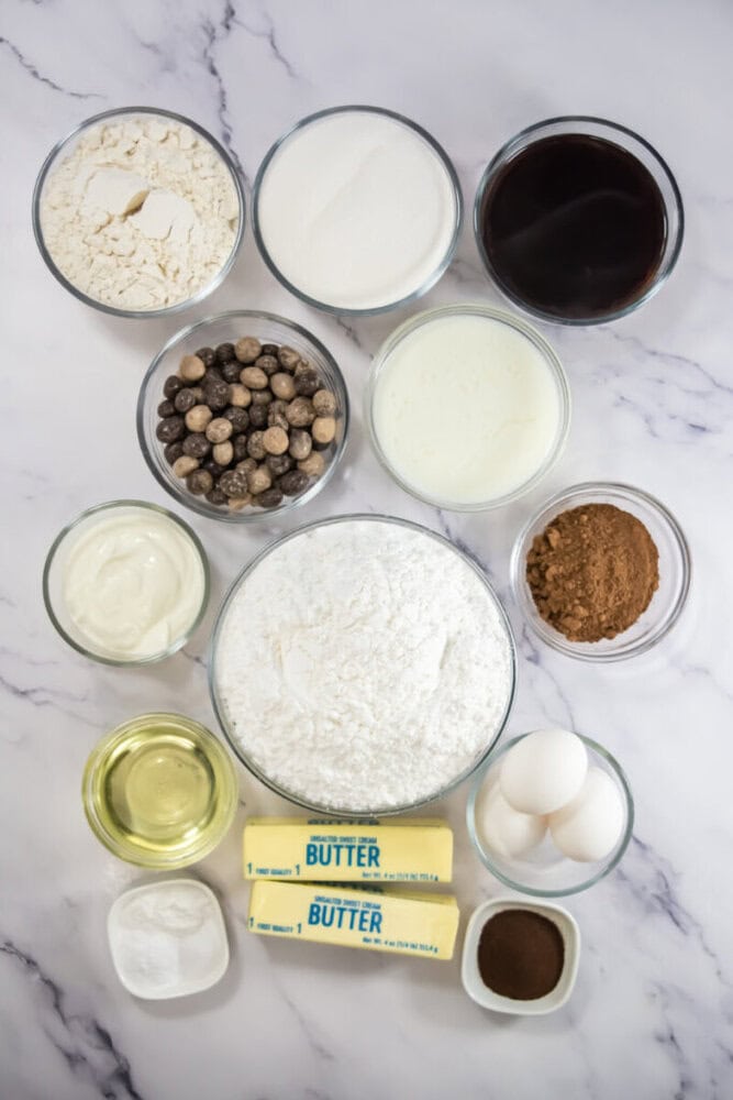 Various baking ingredients, including flour, sugar, cocoa powder, chocolate chips, sour cream, oil, butter sticks, eggs, and bowls of liquid, arranged on a marble surface.
