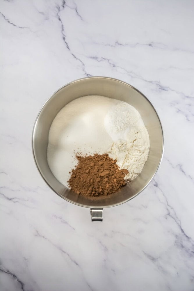 A metal mixing bowl containing cocoa powder, flour, and granulated sugar on a marble surface.