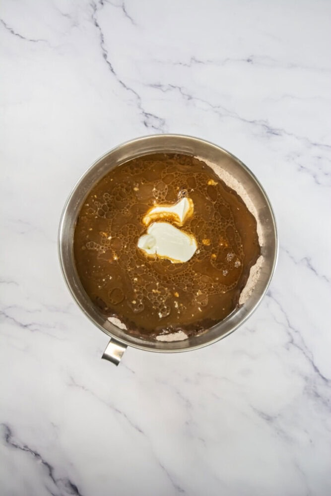 A metal mixing bowl containing brown liquid, oil, and a dollop of white cream, placed on a white marble surface.