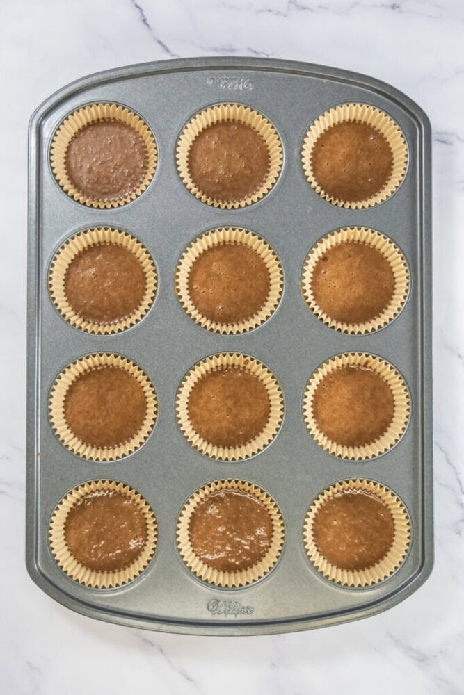 A muffin pan filled with twelve paper cupcake liners, each containing uncooked brown batter, placed on a white marble surface.