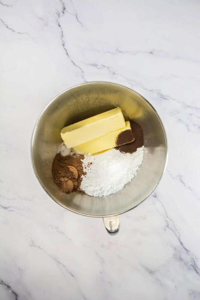 Overhead view of a metal mixing bowl containing two sticks of butter, cocoa powder, powdered sugar, and a small amount of brown liquid on a marble surface.