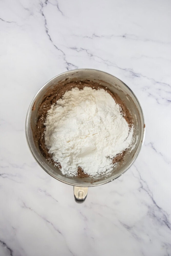 A metal mixing bowl with brown batter and a heap of white flour on top, placed on a white marble surface.