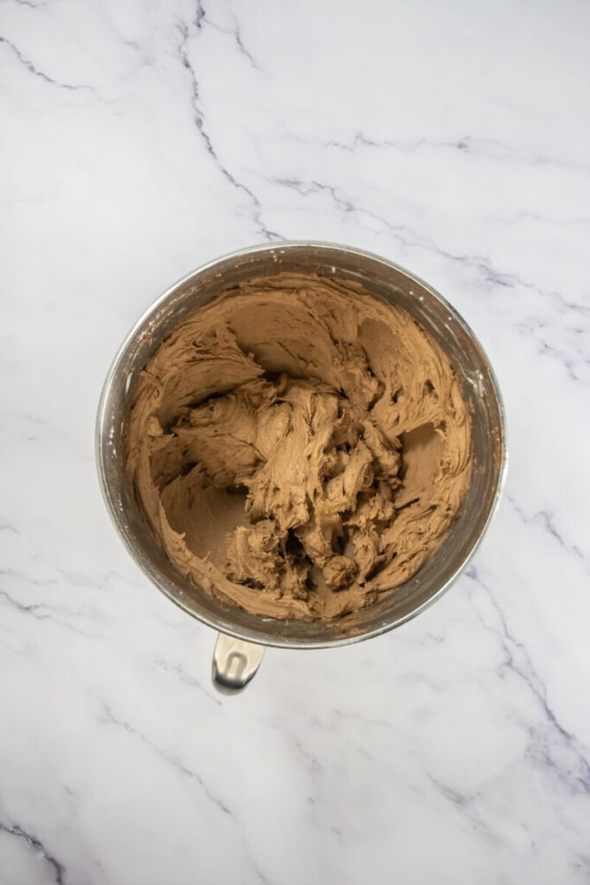 A metal mixing bowl filled with chocolate frosting sits on a white marble surface.