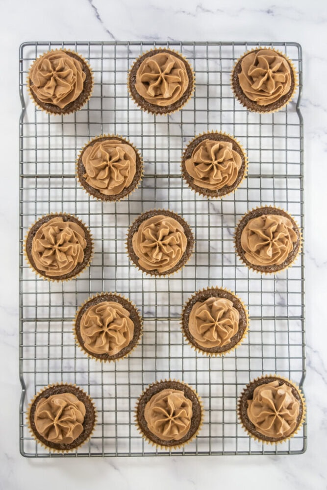 A cooling rack holds fourteen chocolate cupcakes topped with swirled chocolate frosting, viewed from above on a white marble surface.