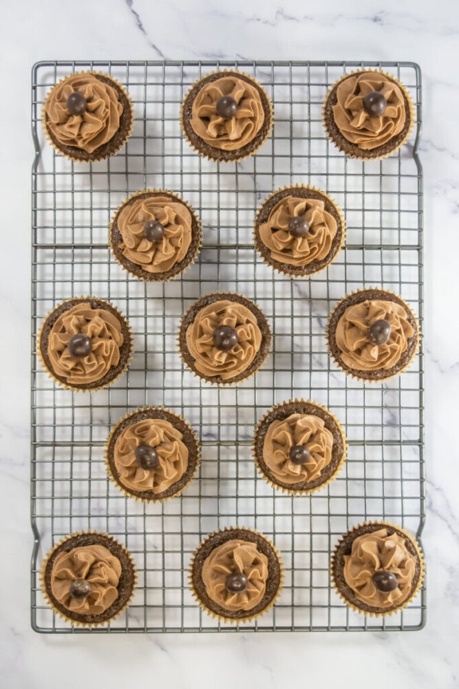 Twelve chocolate cupcakes with swirled chocolate frosting and chocolate candies on top, arranged on a metal cooling rack over a marble surface.