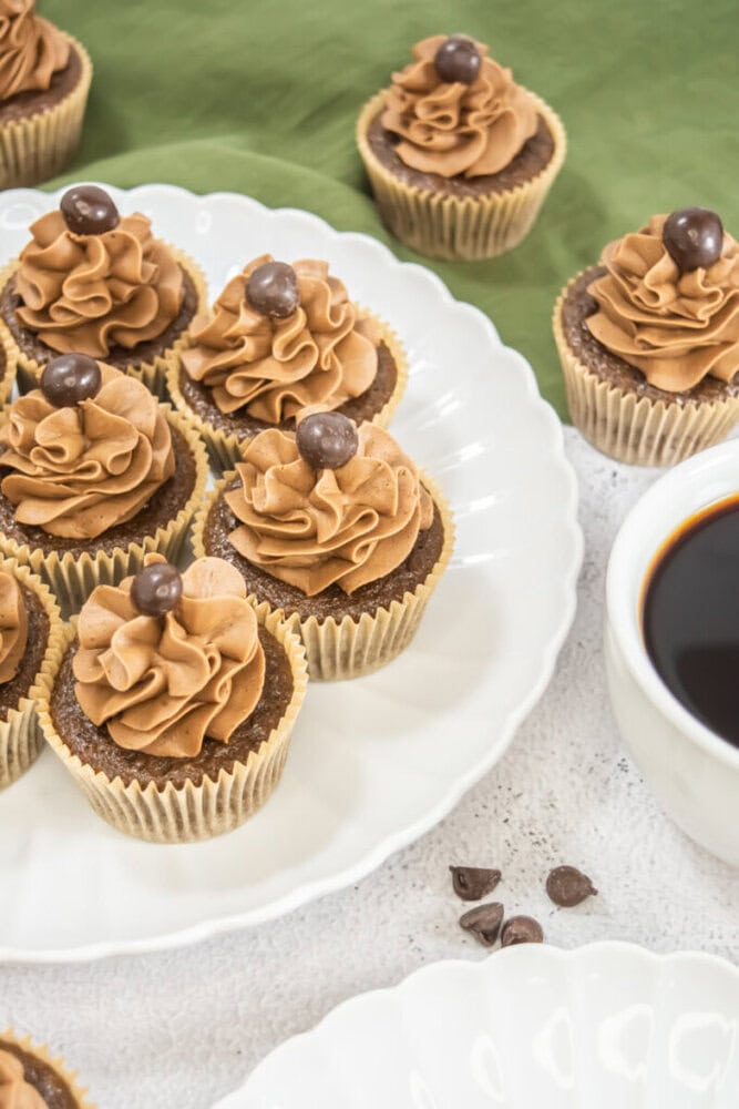 Chocolate cupcakes with swirled mocha frosting and chocolate candy on top are arranged on a white plate next to a cup of black coffee and scattered chocolate chips.
