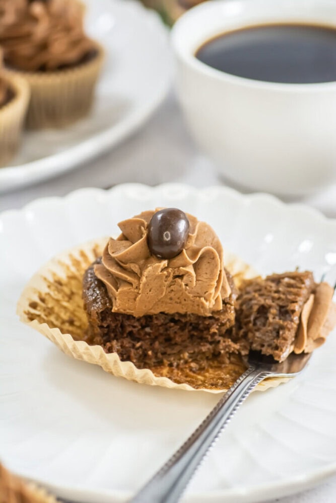 A chocolate cupcake with brown frosting and a coffee bean on top sits on a white plate, partially unwrapped, with a fork holding a bite in front. A cup of black coffee is in the background.