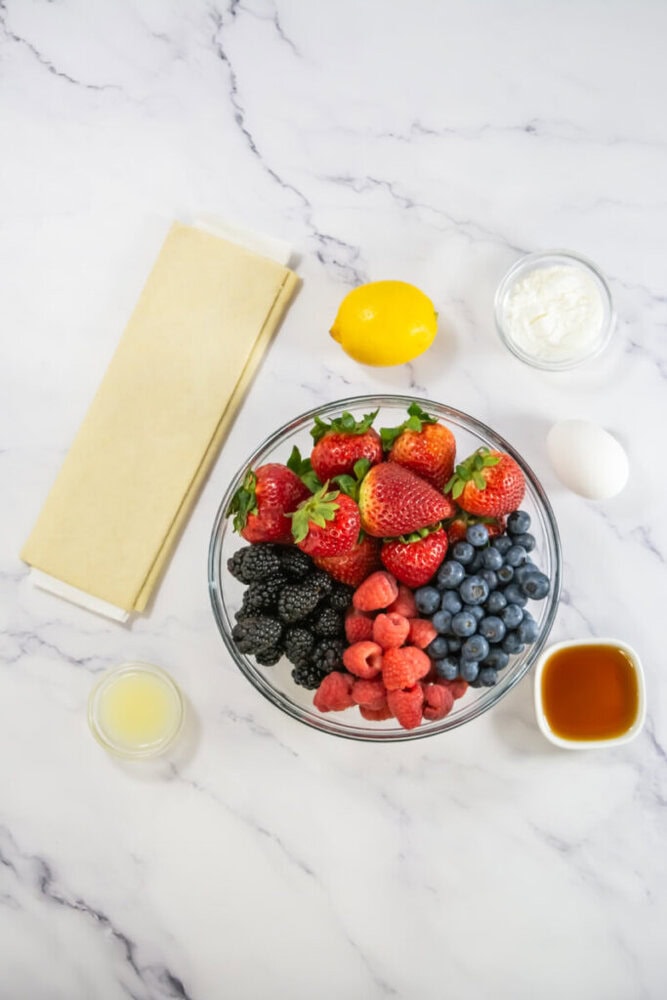 A bowl of fresh strawberries, raspberries, blackberries, and blueberries is surrounded by a lemon, puff pastry sheets, an egg, cream cheese, lemon juice, and syrup on a marble surface.