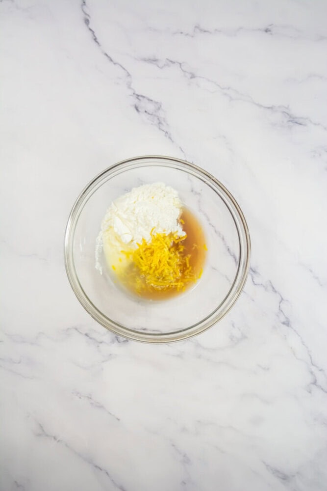 A glass mixing bowl on a marble surface contains flour, a white creamy substance, lemon zest, and a light brown liquid.