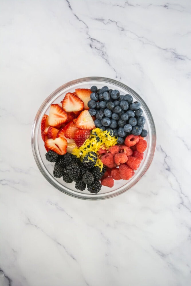 A glass bowl filled with sliced strawberries, blueberries, raspberries, blackberries, and shredded lemon zest on a white marble surface.