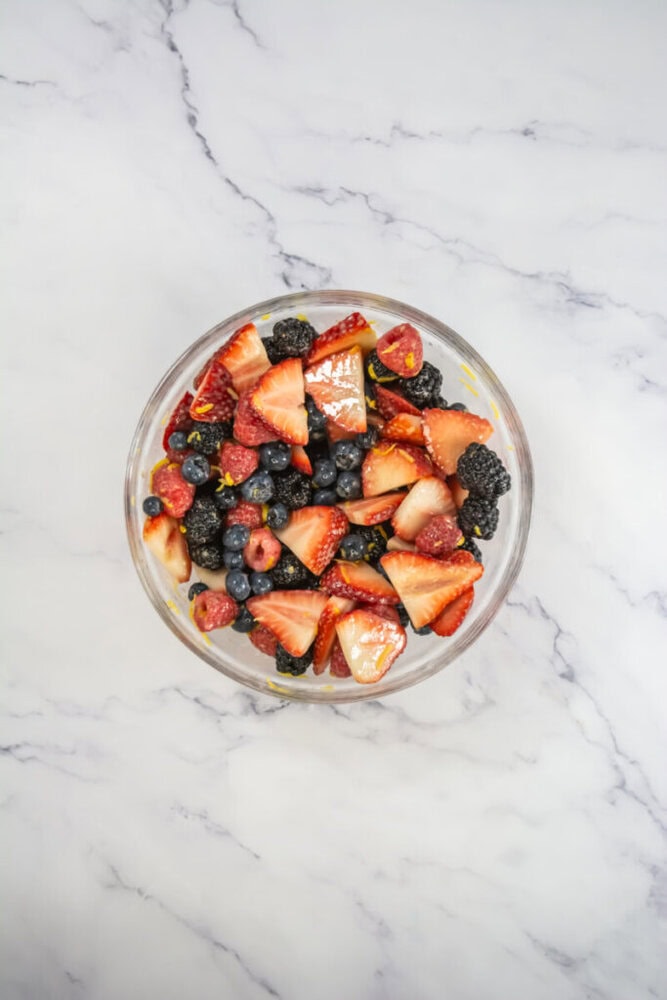 A glass bowl filled with sliced strawberries, blueberries, raspberries, and blackberries on a white marble surface.