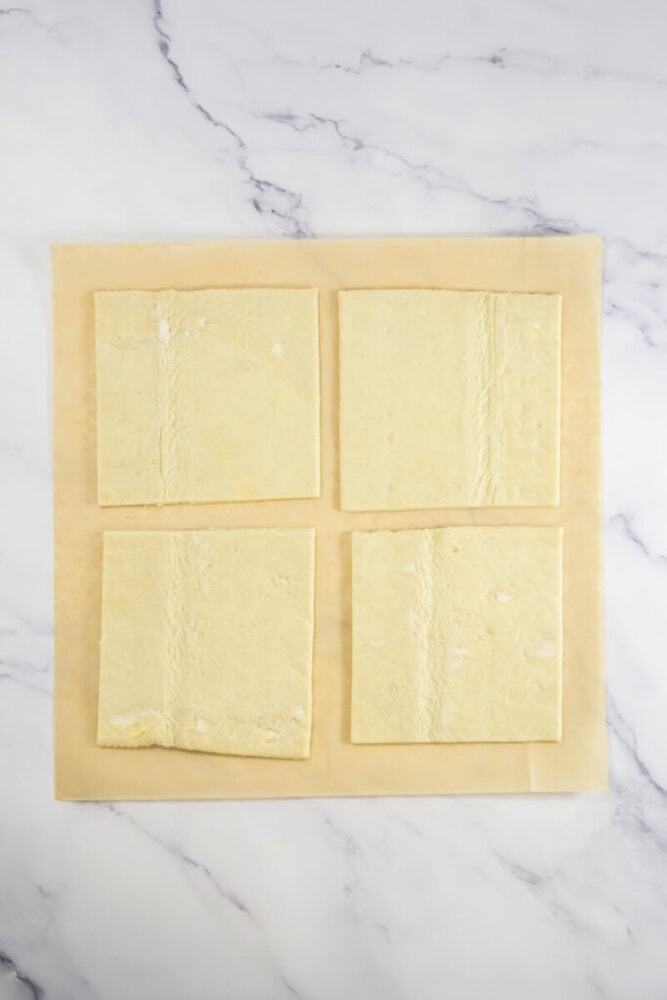 Four square pieces of puff pastry dough are arranged on a sheet of parchment paper on a marble surface.