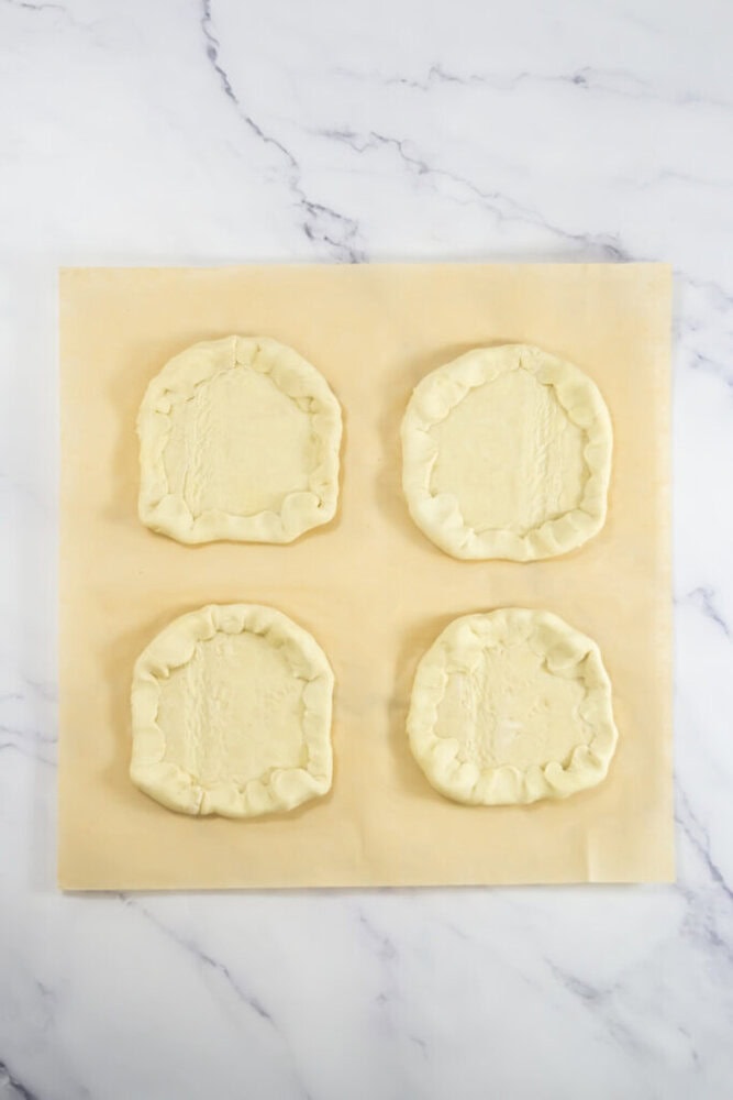 Four unbaked round pastry crusts with folded edges sit on a sheet of parchment paper on a marble countertop.
