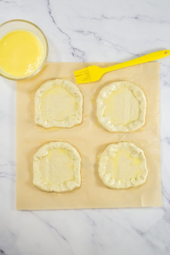 Four unbaked pastry rounds with crimped edges on parchment paper, next to a bowl of egg wash and a yellow pastry brush on a marble surface.