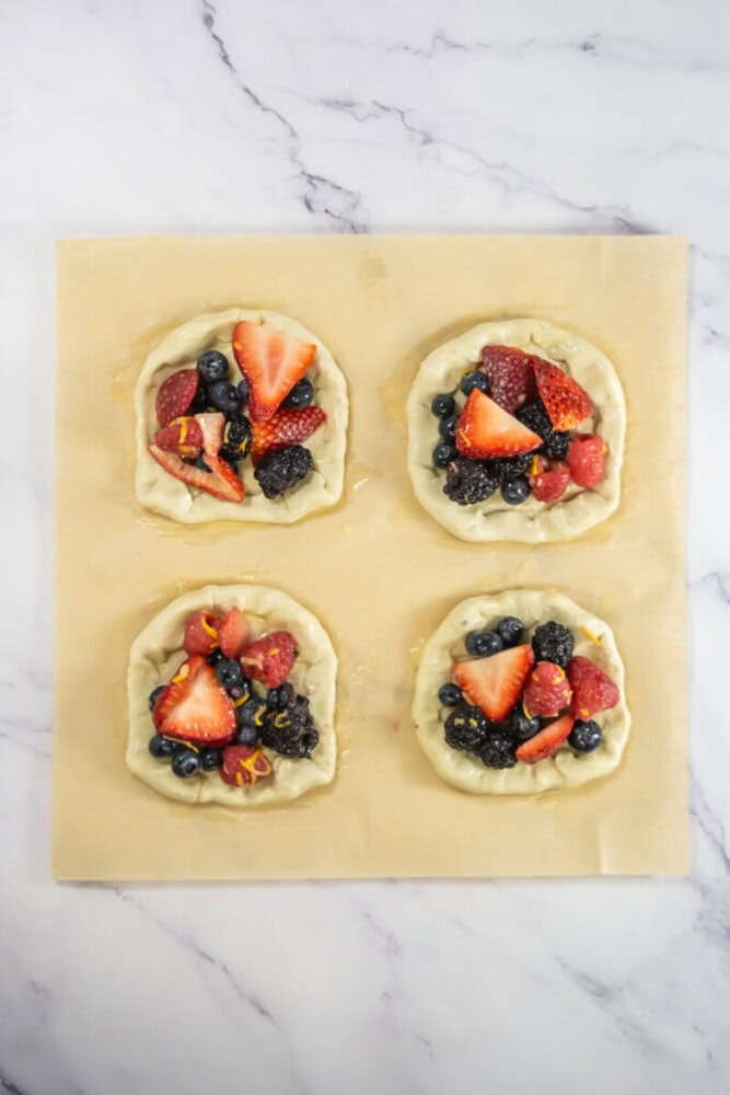 Four unbaked mini galettes filled with mixed berries are arranged on a sheet of parchment paper on a marble surface.