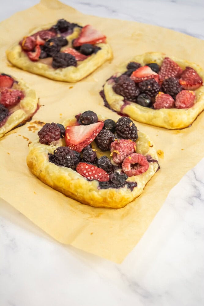 Four square puff pastry tarts topped with assorted berries sit on parchment paper atop a white marble surface.