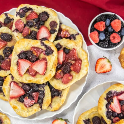 Plates with square pastries topped with strawberries, blueberries, raspberries, and blackberries, alongside a small bowl of assorted fresh berries on a red cloth.