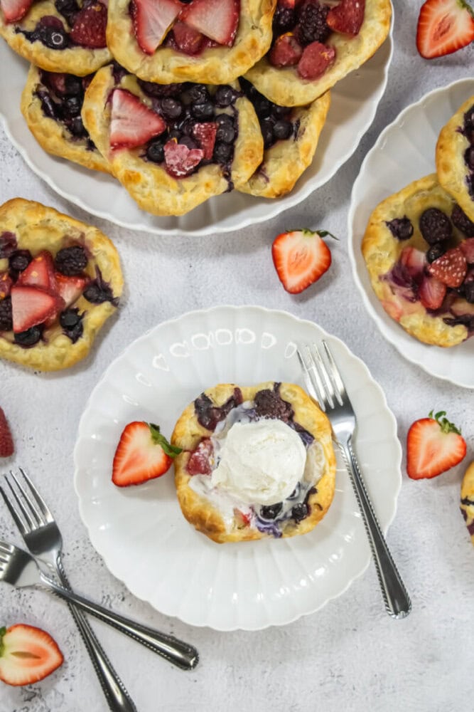 Plates with mixed berry puff pastry tarts, one topped with a scoop of vanilla ice cream, surrounded by fresh strawberries and forks on a light surface.