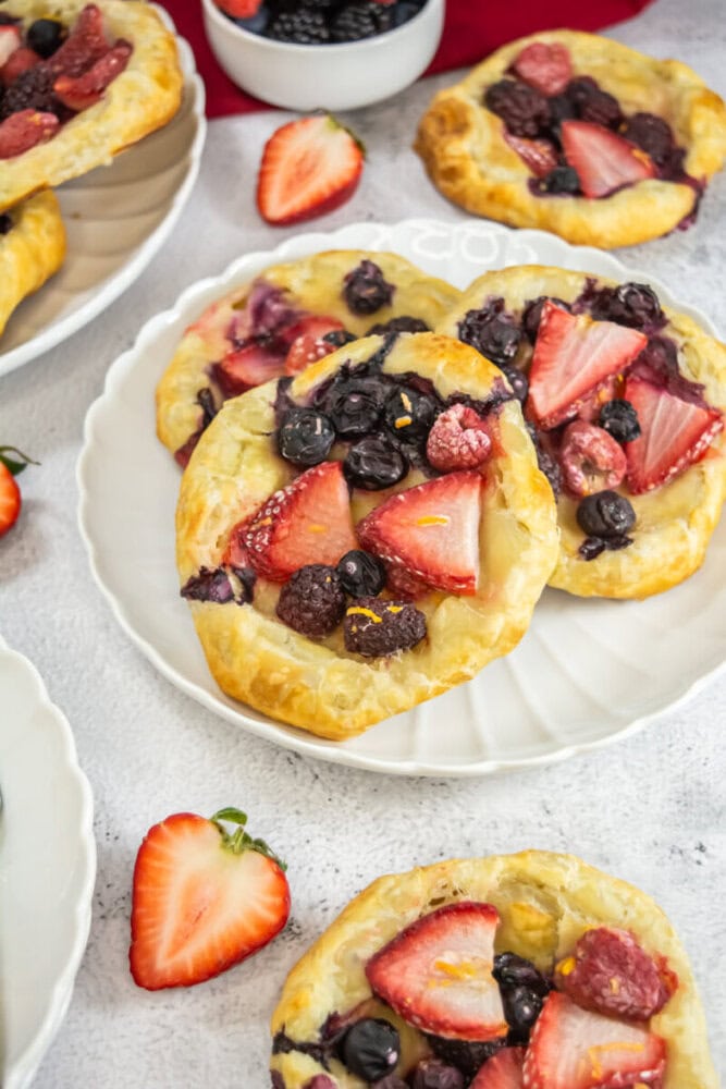 Three mixed berry danishes with strawberries, blueberries, and blackberries sit on a white plate, surrounded by fresh berries on a light surface.