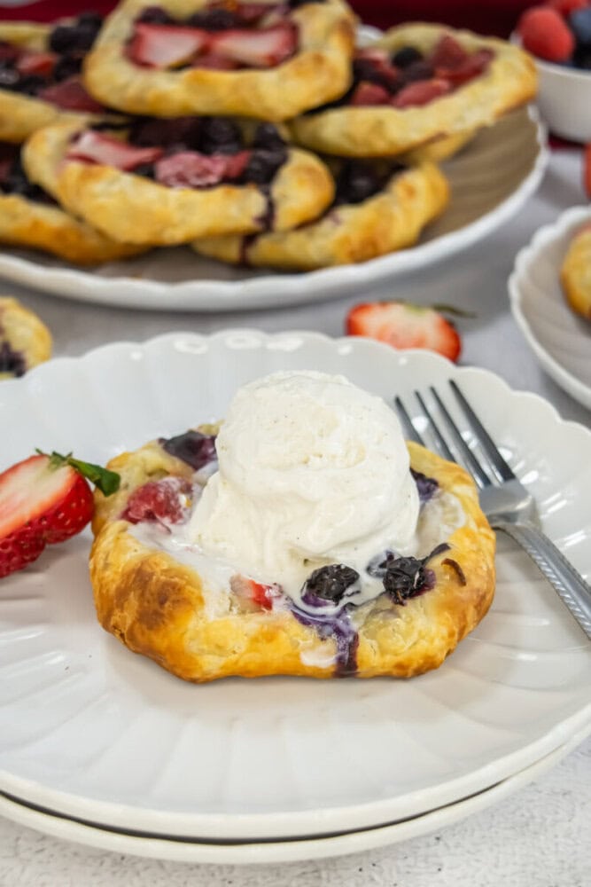 A mixed berry pastry topped with a scoop of vanilla ice cream is served on a white plate with a fork and a sliced strawberry, with more pastries in the background.