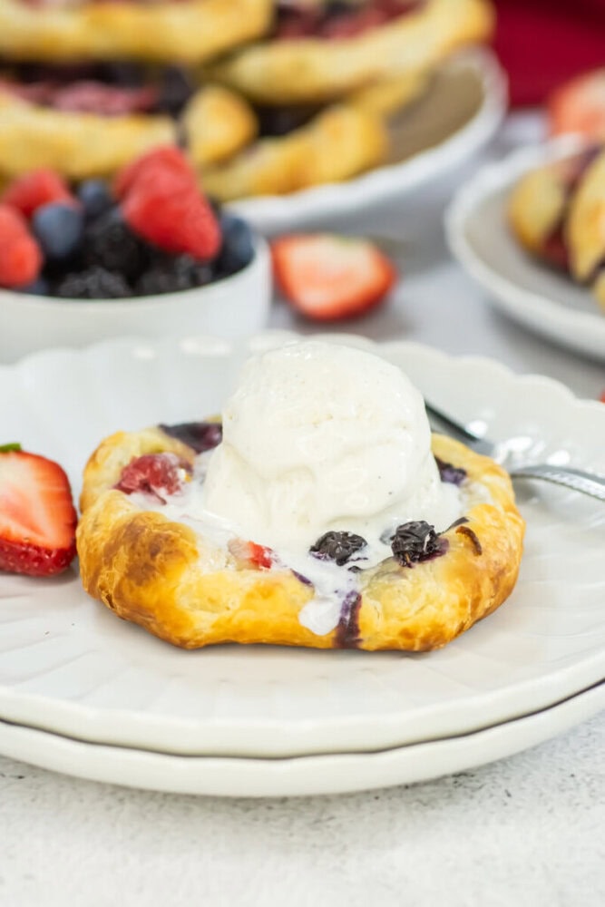 A small fruit tart topped with a scoop of vanilla ice cream on a white plate, with sliced strawberries and mixed berries in the background.
