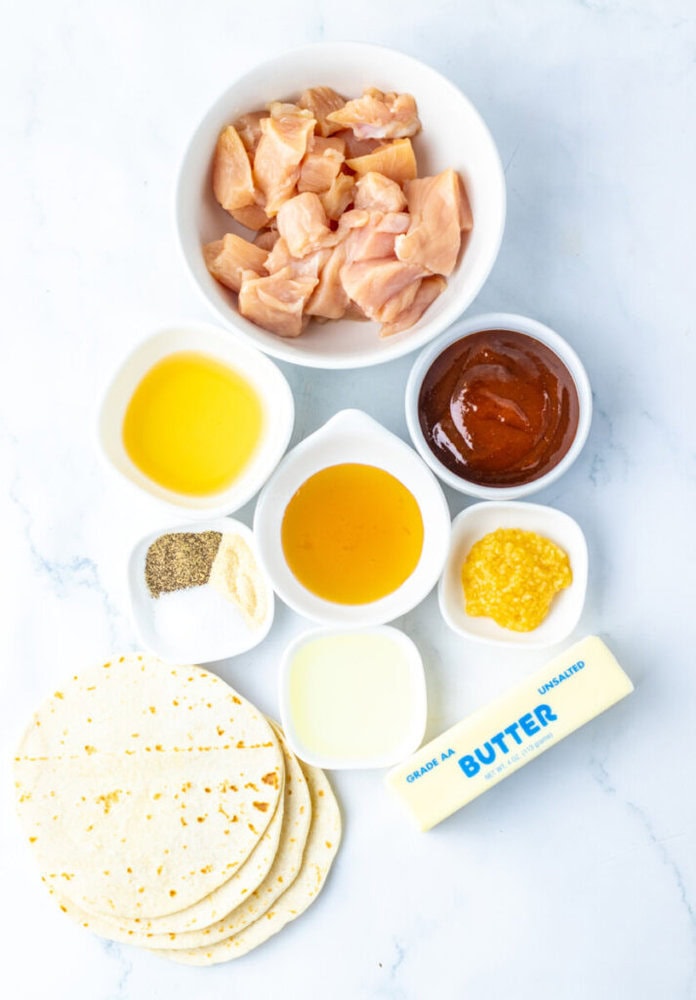 A variety of ingredients including raw diced chicken, barbecue sauce, butter, tortillas, oil, spices, honey, and minced garlic arranged in bowls and containers on a white surface.