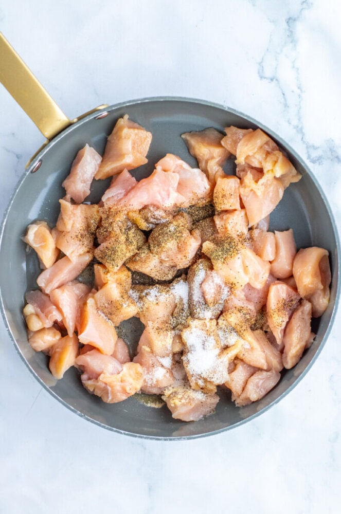 A frying pan with raw, cubed chicken pieces seasoned with salt, pepper, and other spices on a white marble surface.