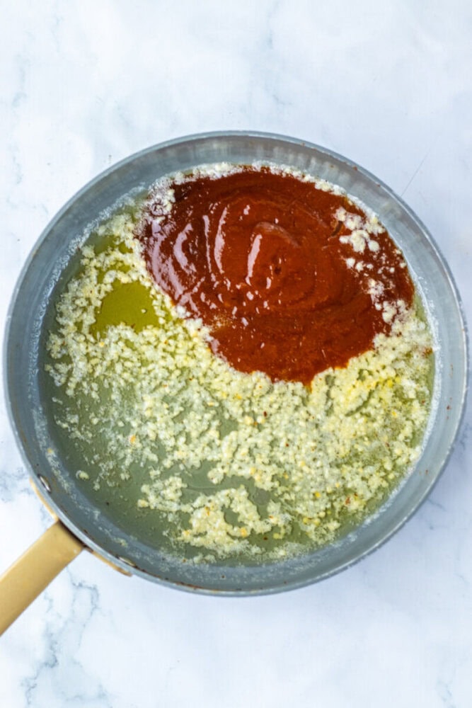 A saucepan containing melted butter, minced garlic, and red hot sauce sits on a white marble surface.