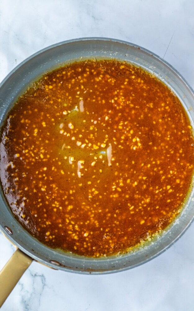 A pan on a marble surface containing a simmering brown sauce with visible minced garlic pieces.