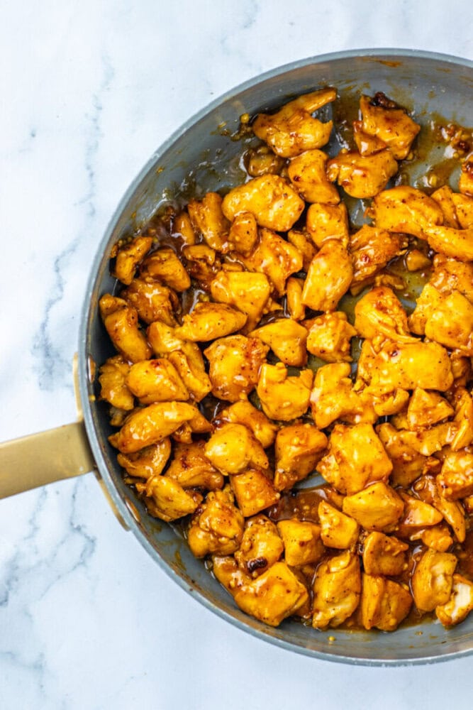 A skillet filled with cooked, diced chicken pieces coated in a golden-brown sauce sits on a white marble surface.