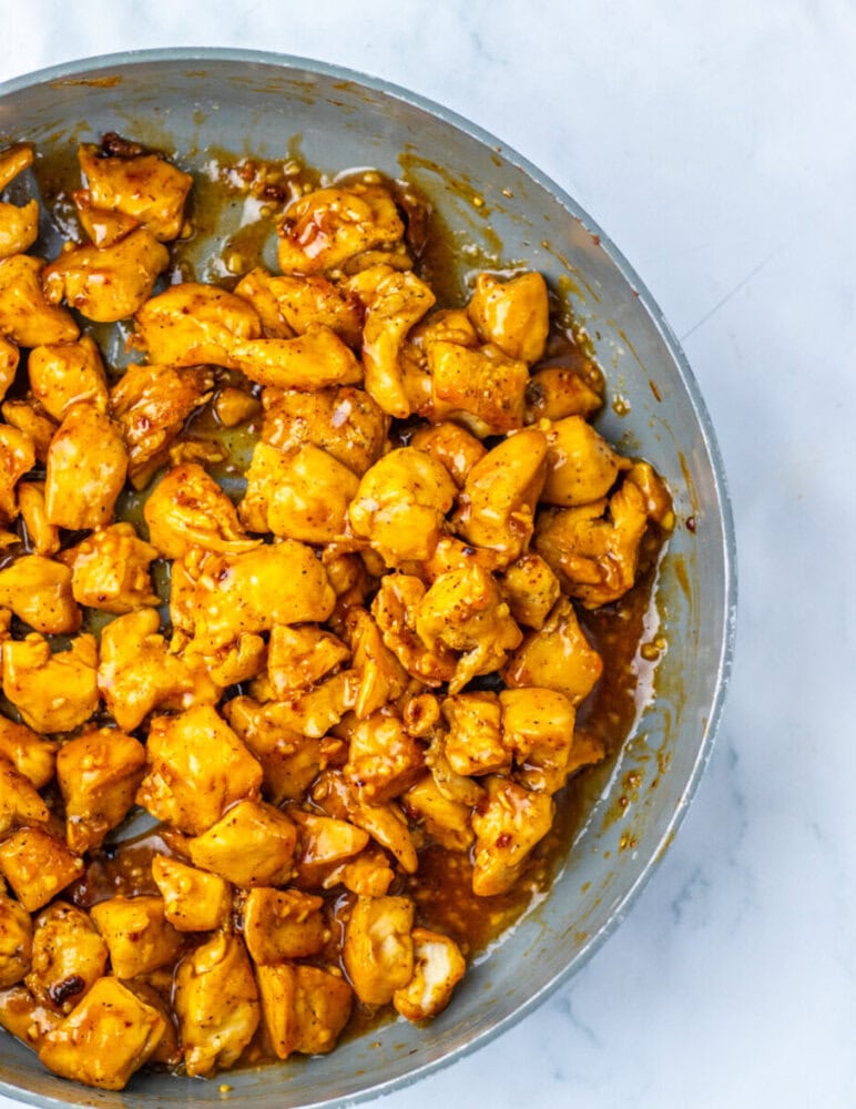 Cooked bite-sized pieces of chicken coated in a glossy, golden-brown sauce in a skillet, photographed from above on a marble surface.