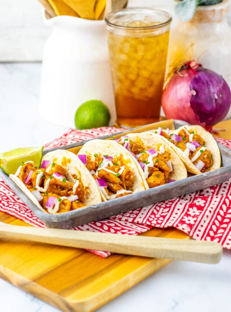 Three tacos filled with seasoned vegetables and topped with chopped red onion and sauce, served on a tray with a lime wedge. Iced tea, a red onion, and a lime are in the background.