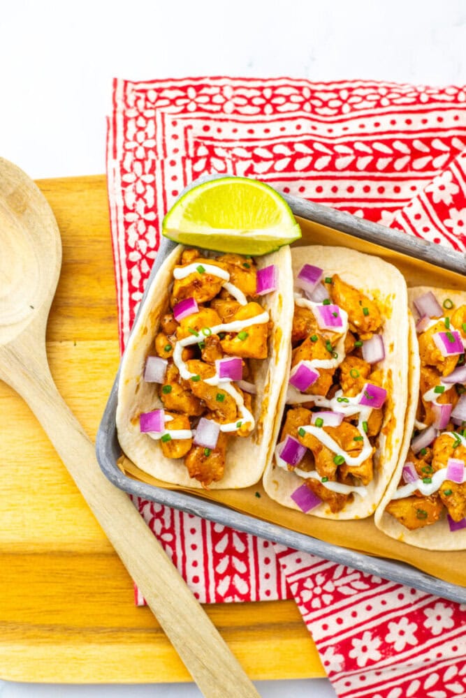 Three tacos filled with diced seasoned chicken, chopped red onion, and cilantro, drizzled with white sauce, served with a lime wedge on a tray, placed on a red patterned napkin.